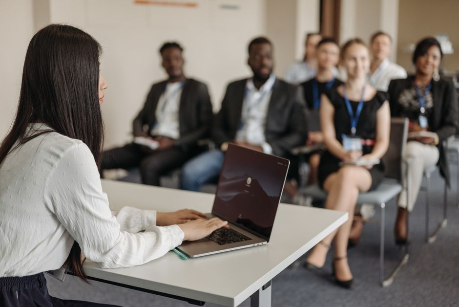Mulher apresentando uma palestra ou sessão de treinamento para um grupo de profissionais em ambiente corporativo, refletindo temas de desenvolvimento e capacitação.