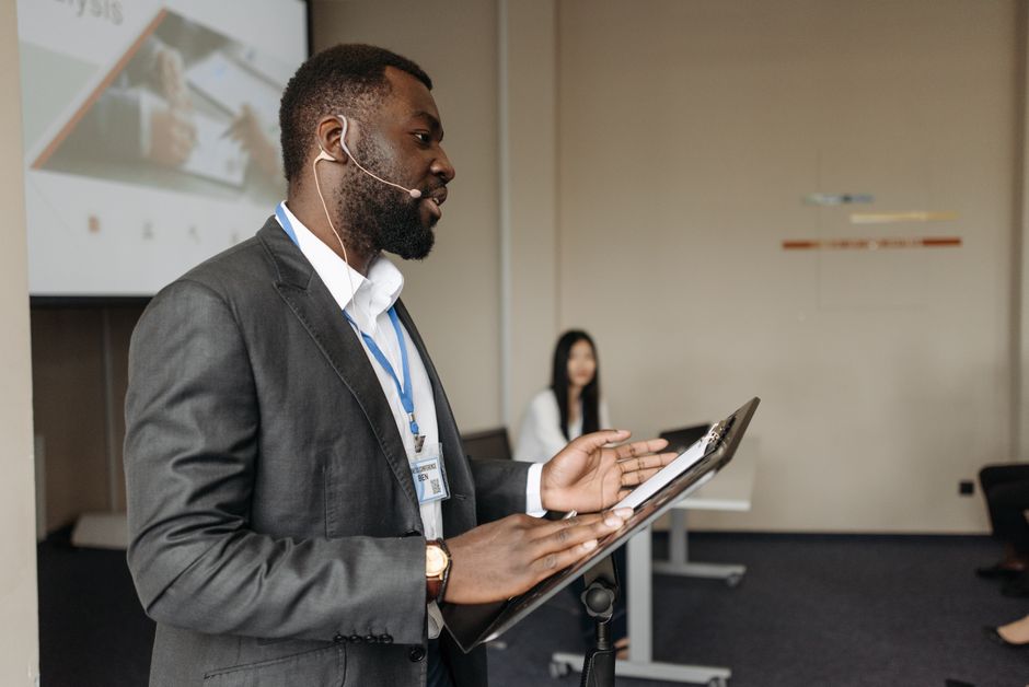 Homem apresentando em uma reunião corporativa, usando tecnologia para discursar em um ambiente de negócios formal.