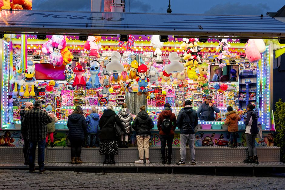 Imagem de uma feira de jogos e prêmios colorida, com pessoas observando os brinquedos e objetos de pelúcia pendurados no estande.