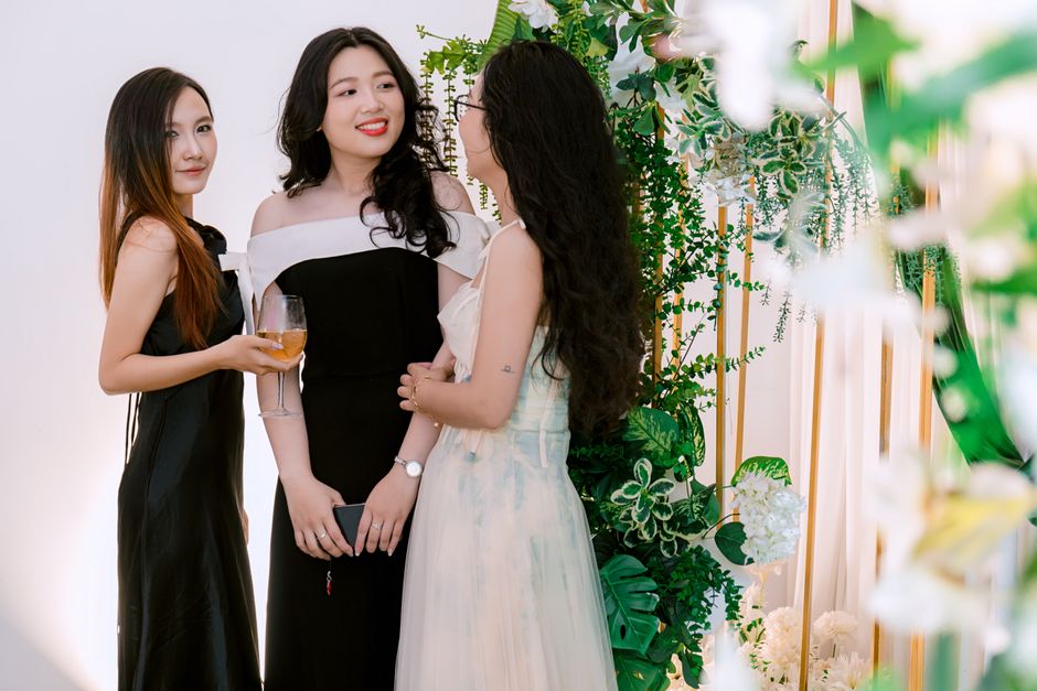 Três mulheres sorrindo e conversando em um evento social elegante, com decoração natural e plantas verdes ao fundo.