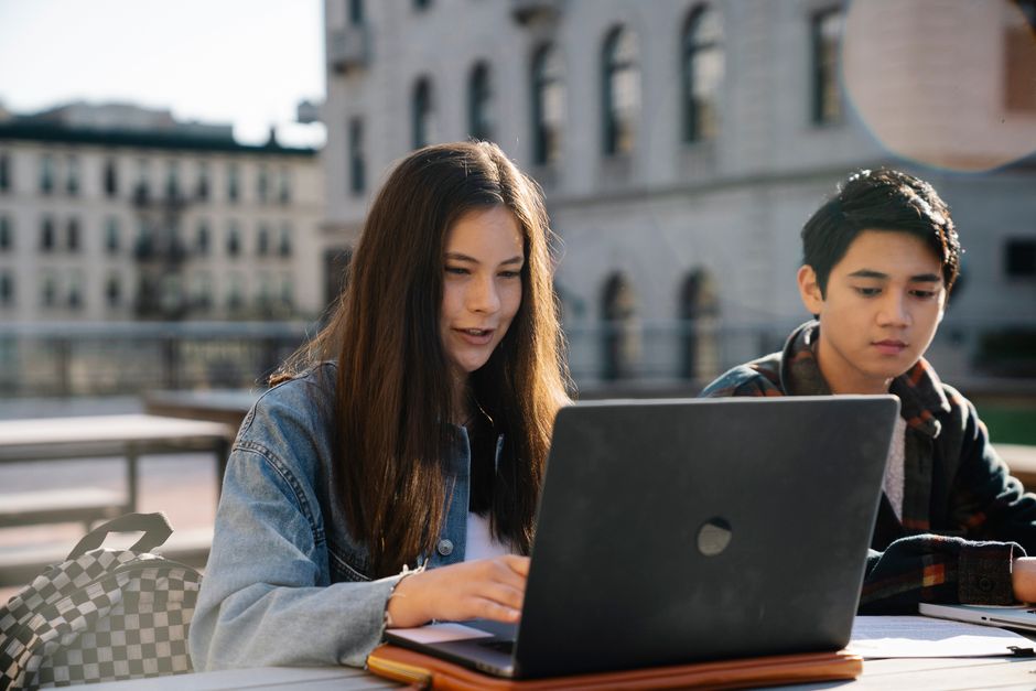 Dois jovens estudando ou trabalhando ao ar livre em um ambiente urbano com foco em tecnologia e educação.