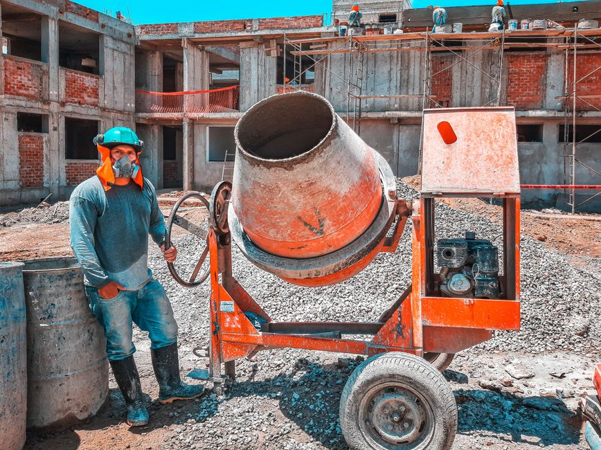 Trabalhador de construção civil operando uma betoneira em uma obra em progresso, com estrutura de edifício ao fundo.