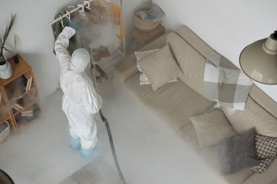 Pessoa usando equipamento de proteção realiza limpeza de descontaminação em ambiente residencial com sofá e ferro de passar roupa ao fundo.