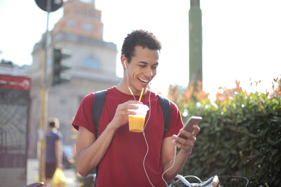Homem jovem sorrindo enquanto escuta música e usa o celular ao ar livre em um ambiente urbano com natureza ao fundo.