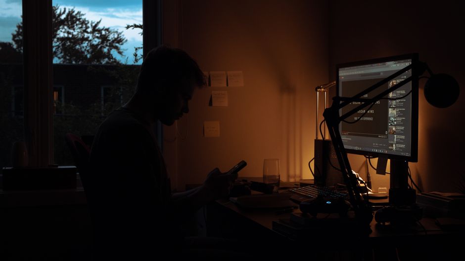 Pessoa trabalhando em um ambiente com iluminação ambiente, usando computador e telefone, refletindo foco na rotina tecnológica e corporativa.