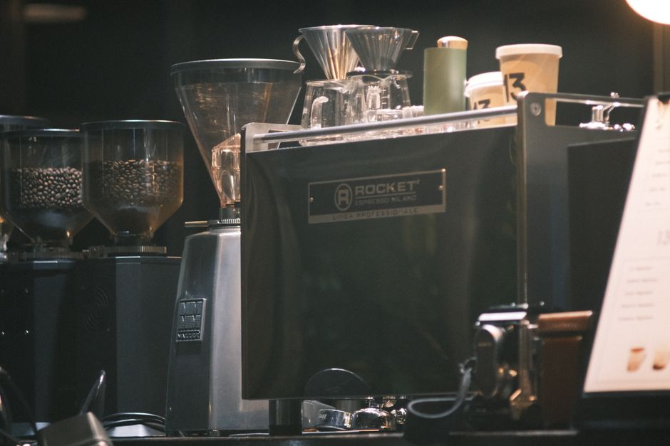 Imagem de uma máquina de café profissional em uma cafeteria moderna, com moedores de café ao fundo, ideal para ilustrar o tema de gastronomia ou cultura de cafés especiais.