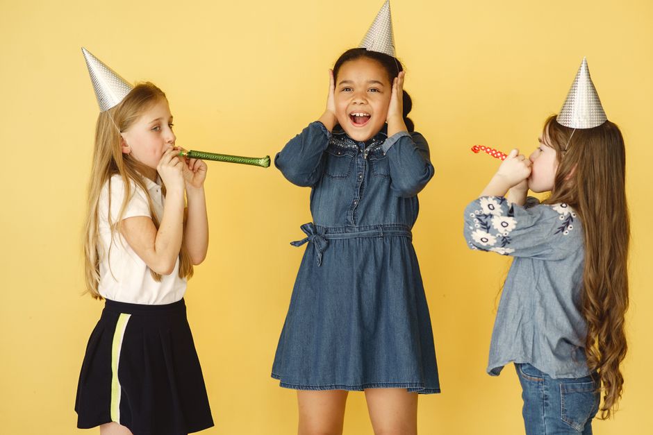 Três crianças comemorando uma festa de aniversário, com as duas meninas brincando de aniversário e a criança no meio expressando surpresa e alegria, em um fundo amarelo.