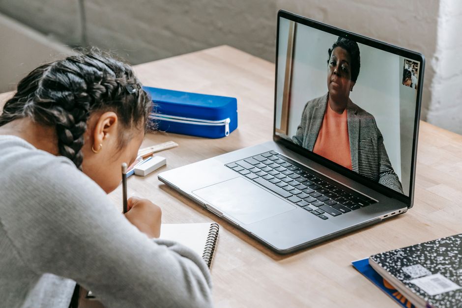 Estudante participando de uma aula ou reunião virtual com orientação de uma professora em vídeo chamada, utilizando computador e materiais de estudo.