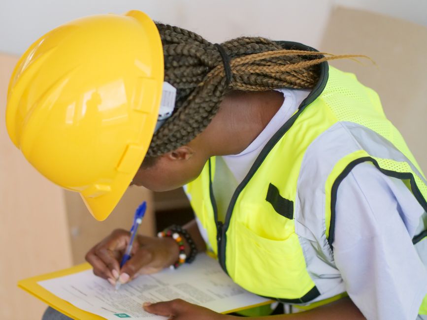 Mulher engenheira fazendo anotações em um ambiente de obra, vestindo capacete de segurança e colete refletivo.