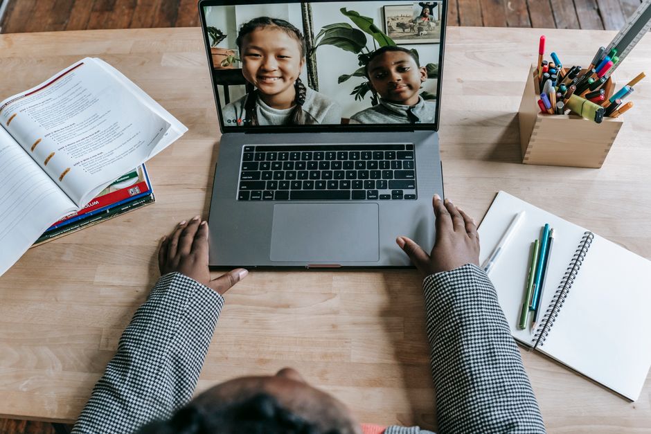 Pessoa participando de uma aula online com duas crianças sorridentes na tela do laptop, em ambiente de estudo em casa.