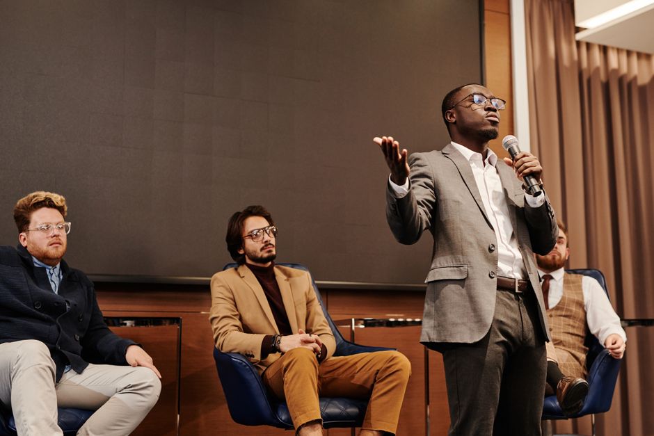 Imagem de um homem negro falando em apresentação ou palestra, com outros homens assistindo e participando do evento, em ambiente formal e profissional.