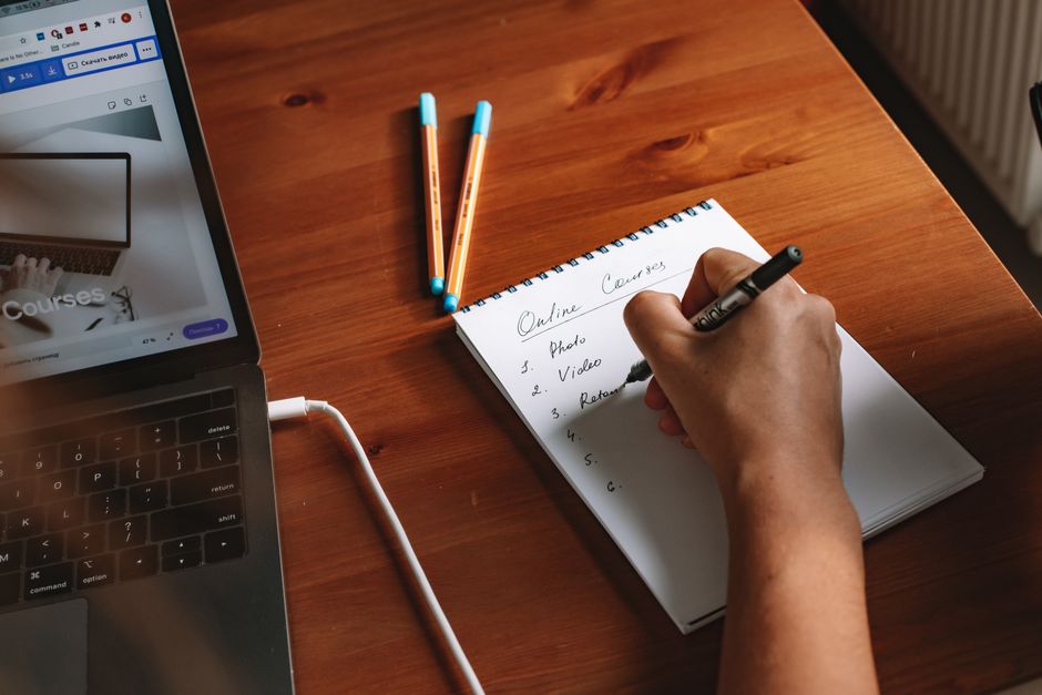 Pessoa fazendo uma lista de tarefas relacionadas a cursos online, com laptop e canetas sobre uma mesa de madeira.