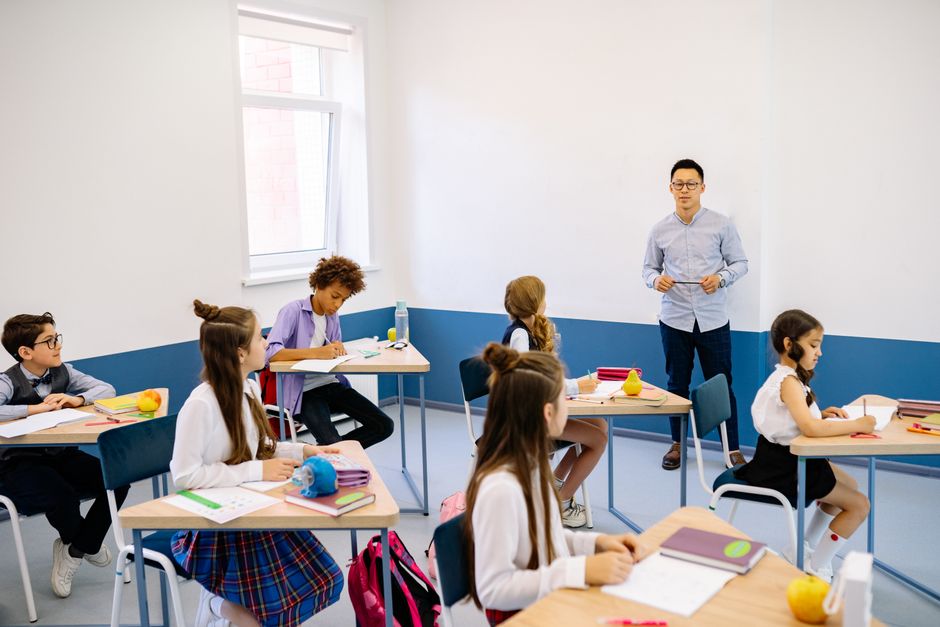 Imagem de uma sala de aula com uma professora diante de alunos, ilustrando um ambiente de ensino e aprendizagem.