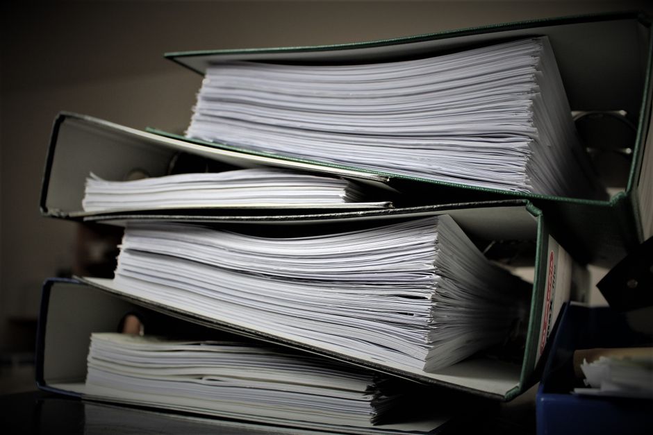 Imagem de várias pilhas de documentos e pastas empilhadas, indicativa de volume de trabalho ou burocracia administrativa.