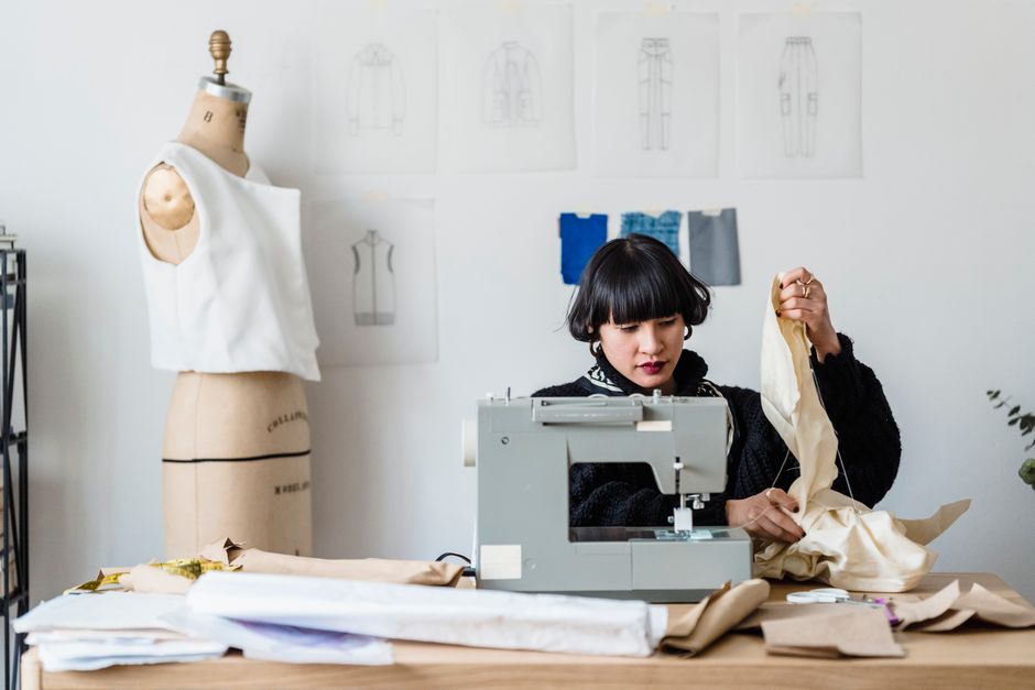 Mulher costurando roupas em um ateliê de moda, cercada por desenhos de modelos e tecidos, focada no trabalho de design e confecção de roupas.