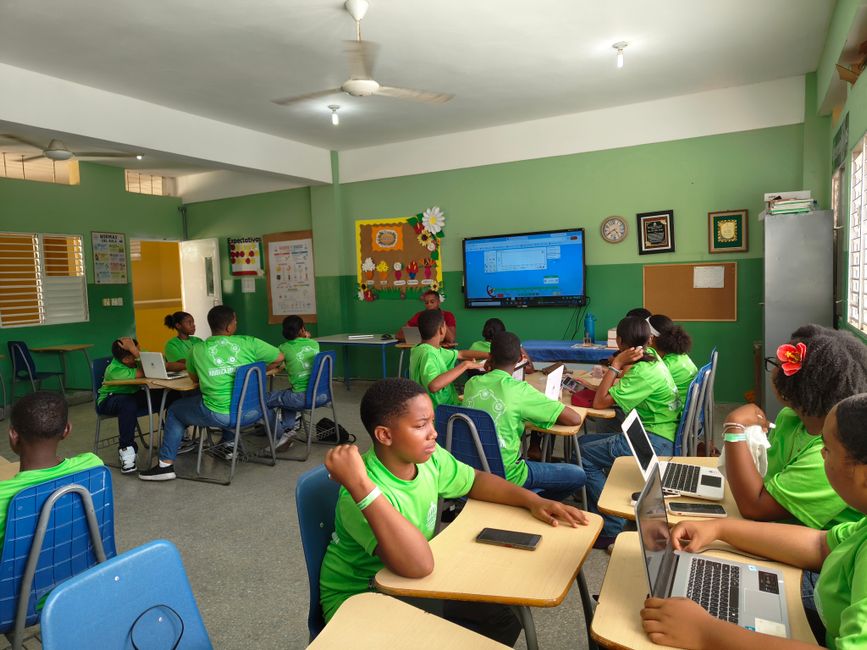 Turma de crianças em sala de aula de informática, participando de atividade educacional com computadores e uma tela digital ao fundo.