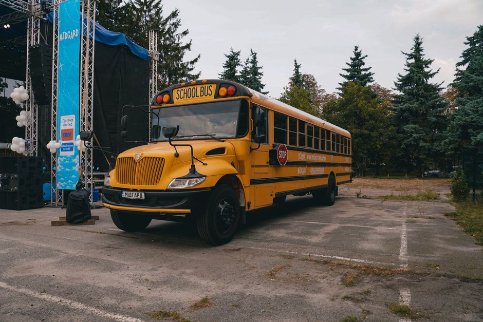 Ônibus escolar amarelo estacionado em área ao ar livre com cenário de árvores e palco ao fundo, indicado para transporte de estudantes e eventos educativos.