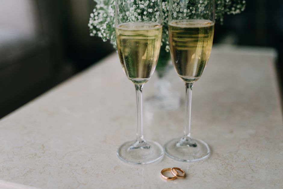 Duas taças de espumante em uma comemoração, com alianças de casamento ao final da mesa, simbolizando celebração e união.
