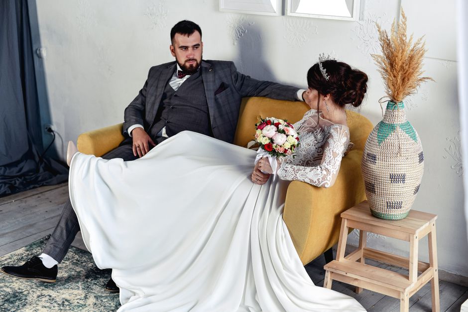 Momento romântico de um casamento, com o noivo sentado na mesa e a noiva segurando um buquê de flores, em um ambiente acolhedor.