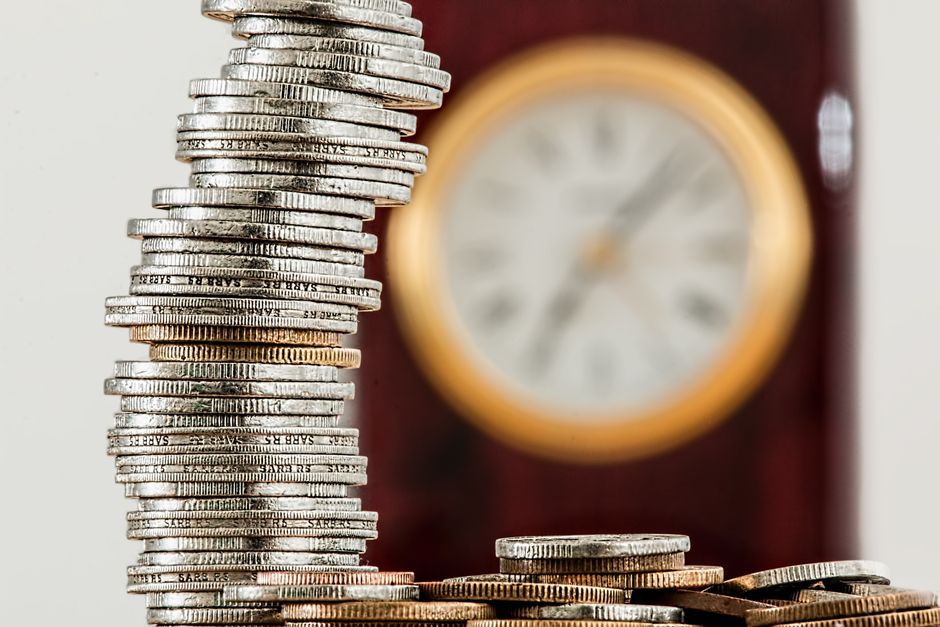Imagem de uma pirâmide de moedas de diferentes tamanhos, simbolizando conceitos de economia e finanças, com um relógio ao fundo indicando gestão de tempo ou planejamento financeiro.