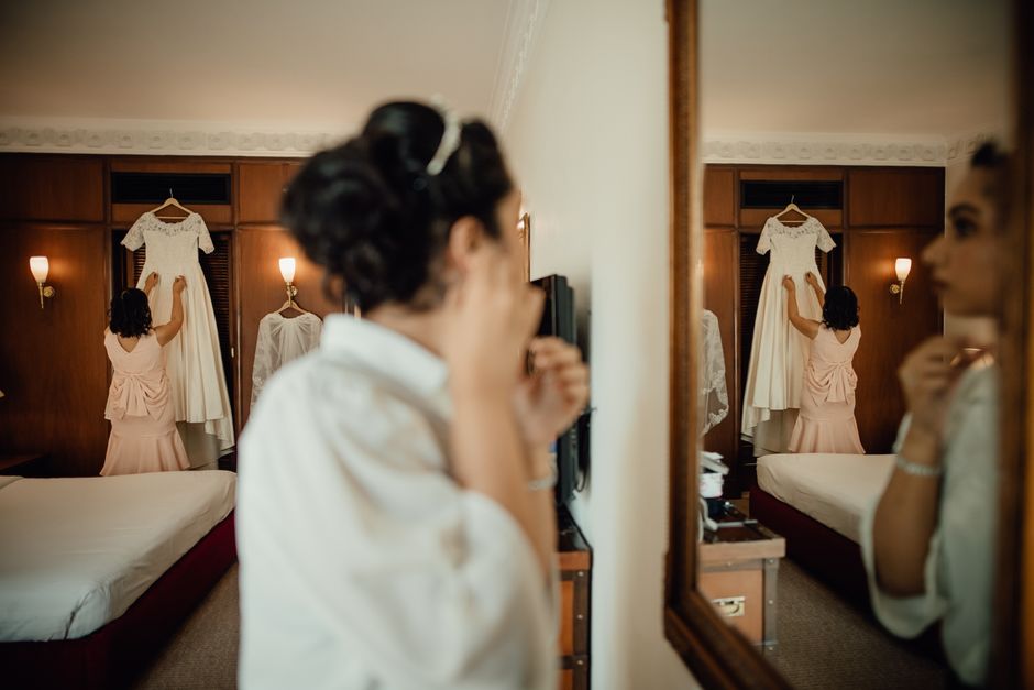 Mulher se arrumando para um evento especial, refletida em um espelho na sala de um hotel ou casa, com vestido de casamento pendurado ao fundo.