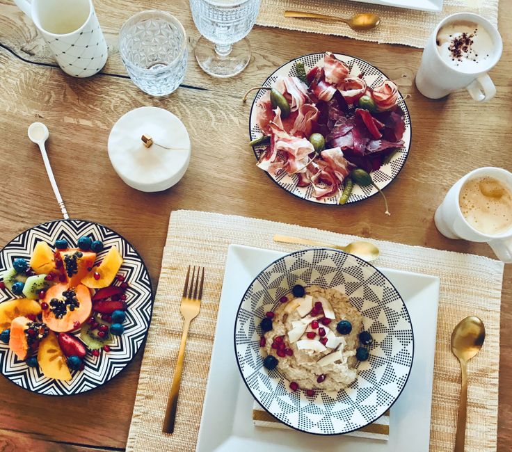 Mesa de café da manhã saudável com frutas, queijos, presunto, cappuccino e utensílios de ouro, ideal para um momento de lazer ou pausa no dia.