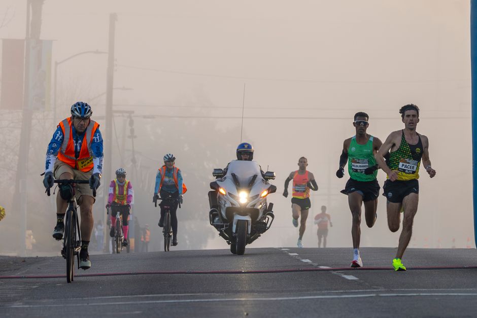 Atletas participando de uma corrida urbana sob neblina, com motociclista de apoio ao fundo.