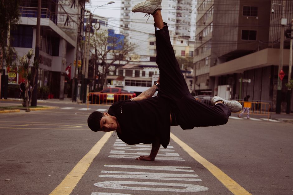 Pessoa fazendo uma manobra de break dance na rua de uma cidade urbana durante o dia, com edifícios e trânsito ao fundo.