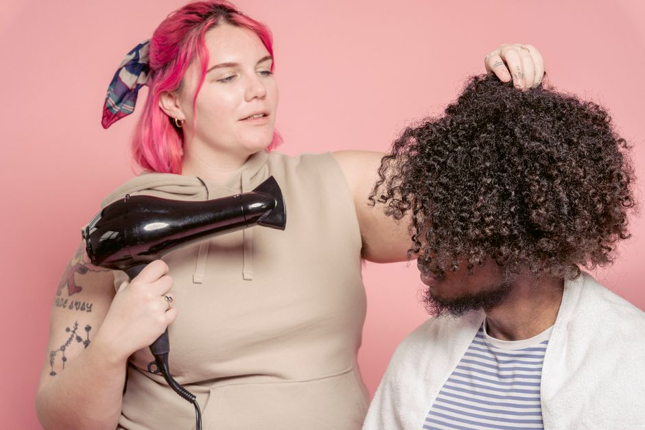 Retrato de uma mulher usando secador de cabelo próximo à cabeça de um homem, em um contexto de cuidados pessoais ou beleza, com fundo rosa claro.