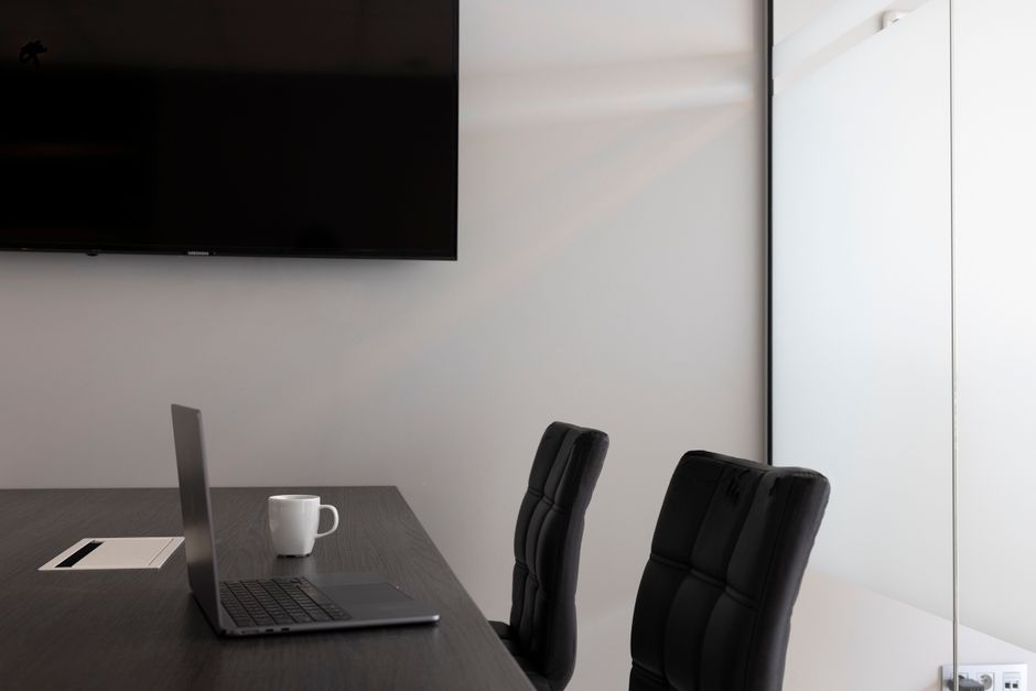 Imagem de uma sala de reunião moderna e minimalista com uma mesa de escritório, cadeiras ergonômicas, um computador portátil, uma xícara e uma tela grande na parede, ideal para artigos sobre gestão e ambiente corporativo.