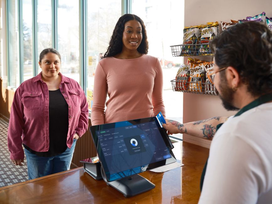 Mulher fazendo pagamento com cartão em uma loja, enquanto uma cliente observa ao fundo, destacando o tema de atendimento ao cliente e uso de tecnologia no comércio.