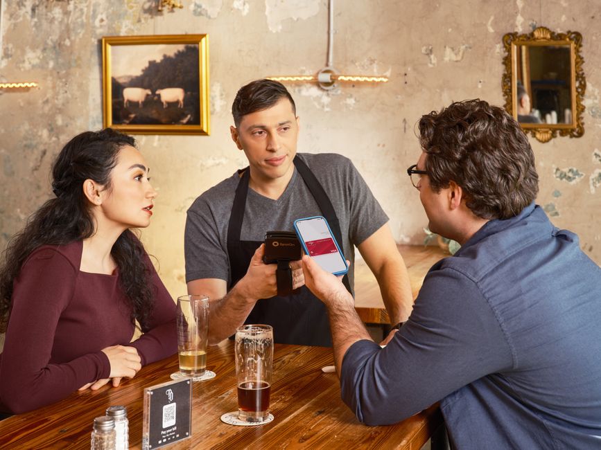 Um casal fazendo pagamento em um restaurante enquanto um garçom registra a transação com um dispositivo móvel, ilustrando o tema de pagamentos e tecnologia na gastronomia.