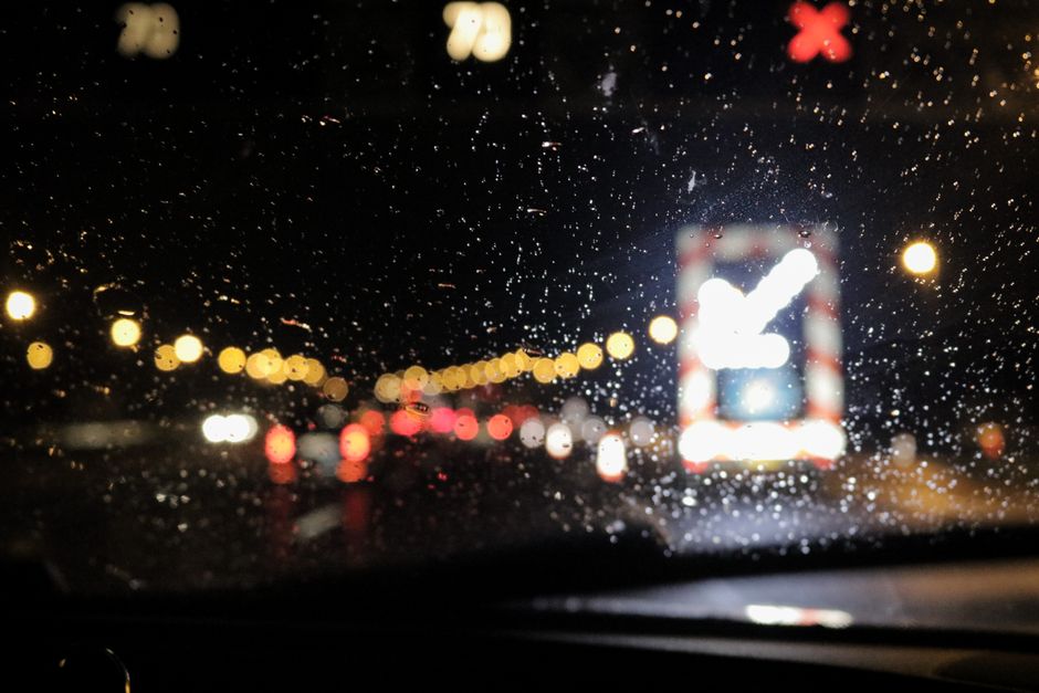 Imagem de uma estrada à noite vista através de uma janela com chuva, criando um efeito de desfoque e luzes difusas.