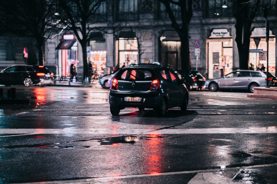 Cena urbana noturna na rua com carros e lojas iluminadas ao fundo, destacando o movimento e a atmosfera de cidade à noite.