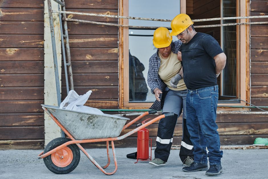 Duas pessoas trabalhando em uma construção residencial, usando capacetes de segurança e verificando algo próximo a uma betoneira, em um ambiente de obra civil.