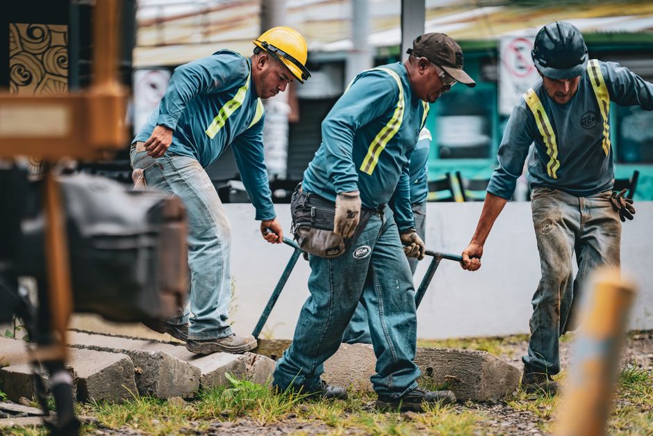 Três trabalhadores construindo uma calçada, demonstrando esforço físico e trabalho manual em ambiente externo.