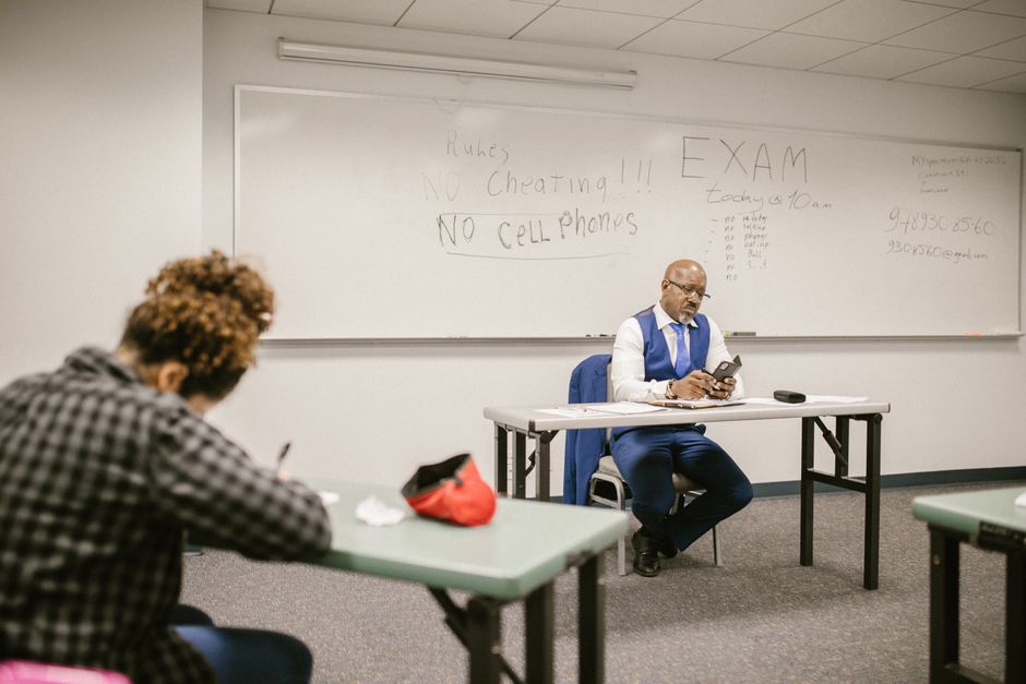 Professor em sala de aula atento ao celular durante uma avaliação, com avisos de regras na lousa ao fundo.