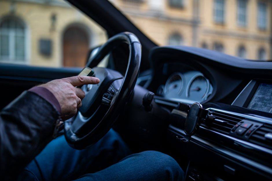 Imagem de um motorista ao volante de um veículo urbano, com foco no painel e na interação com o aparelho móvel, capturada em ambiente de cidade.