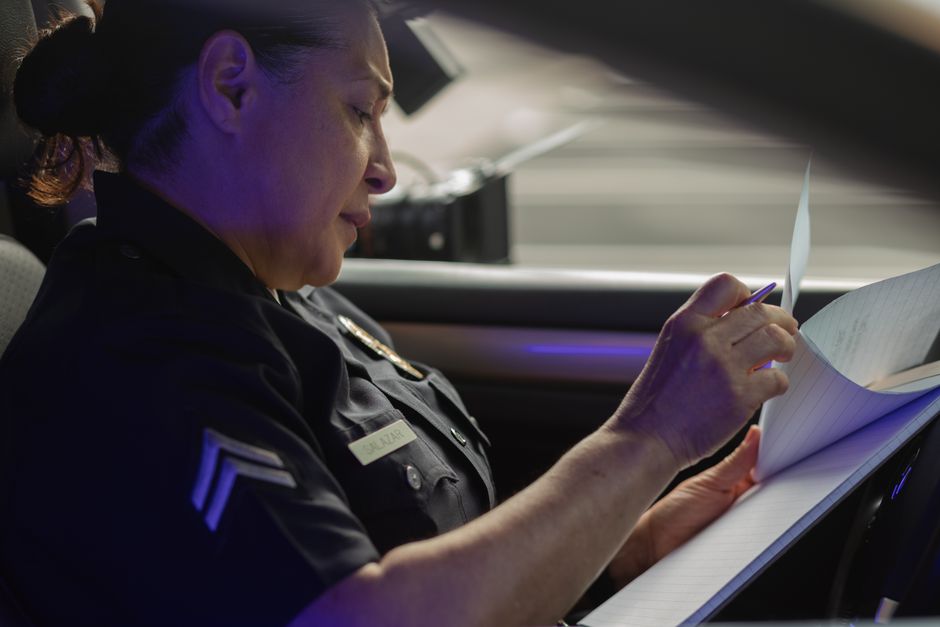 Policial revisando registros ou relatórios dentro de um carro de patrulha, simbolizando investigação ou patrulhamento policial.