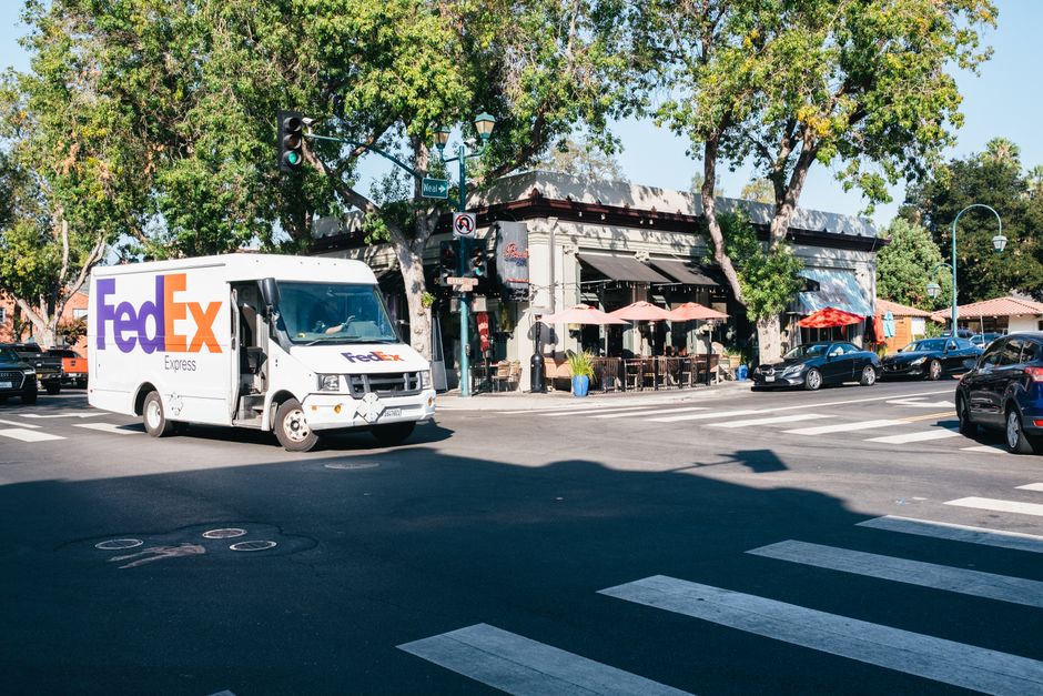 Imagem de uma rua movimentada com um caminhão de entrega da FedEx em frente a um café ao ar livre, com árvores e veículos estacionados ao redor, ilustrando o tema de logística urbana e entregas rápidas.