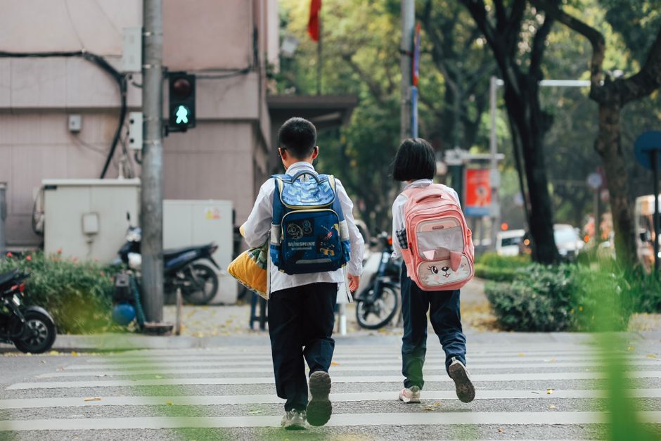 Crianças indo para a escola atravessando uma faixa de pedestres em uma rua arborizada, representando o início do dia escolar.