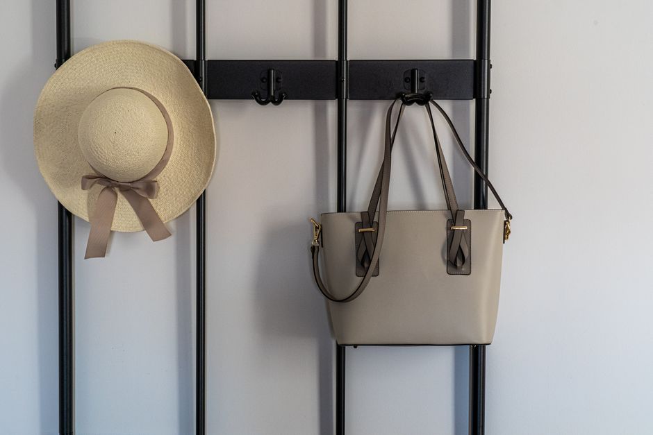 Imagem de uma parede com cabideiro onde estão pendurados um chapéu de brim com fita, uma bolsa de couro e um cabide, sugerindo organização e estilo na entrada do ambiente.