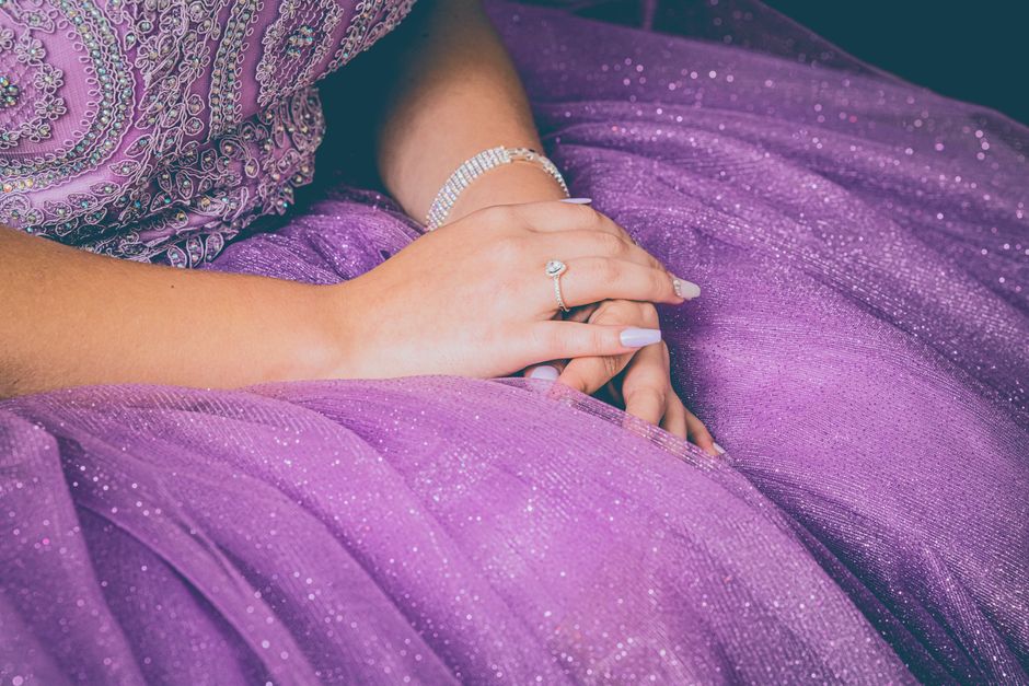 Mão de uma mulher com joias descansando em um vestido roxo brilhante, transmitindo elegância e celebração.
