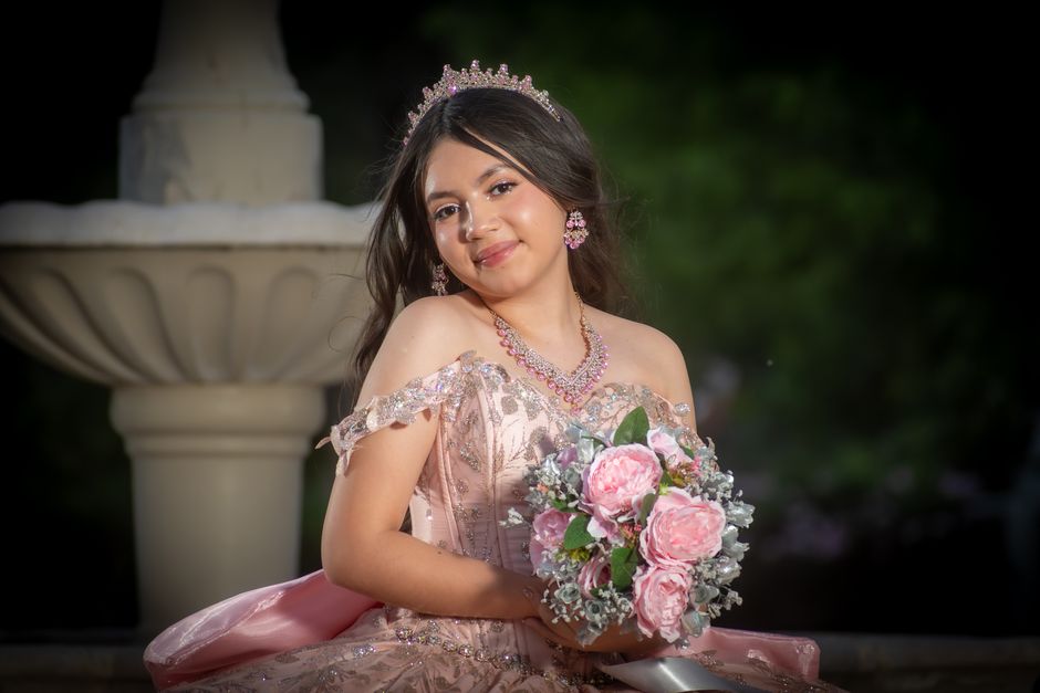 Menina vestida de rosa com tiara e joias, segurando um buquê de flores, em um ambiente externo elegante, ideal para comemoração de aniversário ou ocasião especial.