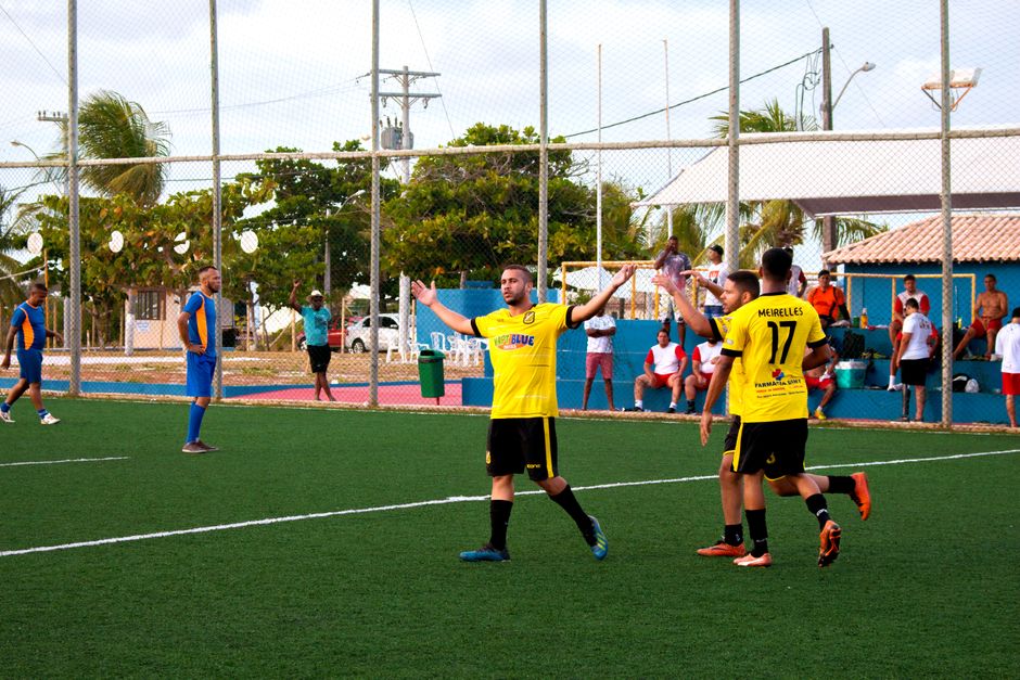 Jogadores comemorando um gol ou jogada importante durante uma partida de futebol em um campo ao ar livre, com espectadores ao fundo e clima descontraído.