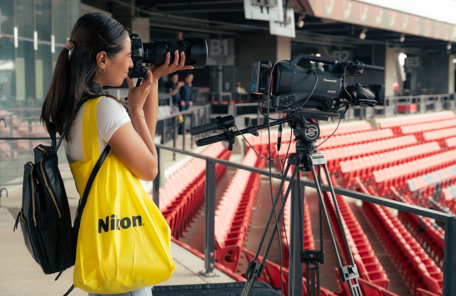 Jornalista ou videomaker capturando imagens em um evento esportivo ao ar livre, com estruturas de estádio ao fundo.