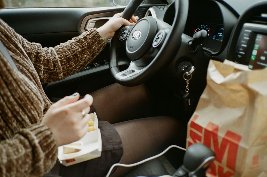 Pessoa dirigindo um carro enquanto faz uma pausa para comer fast food durante uma viagem.