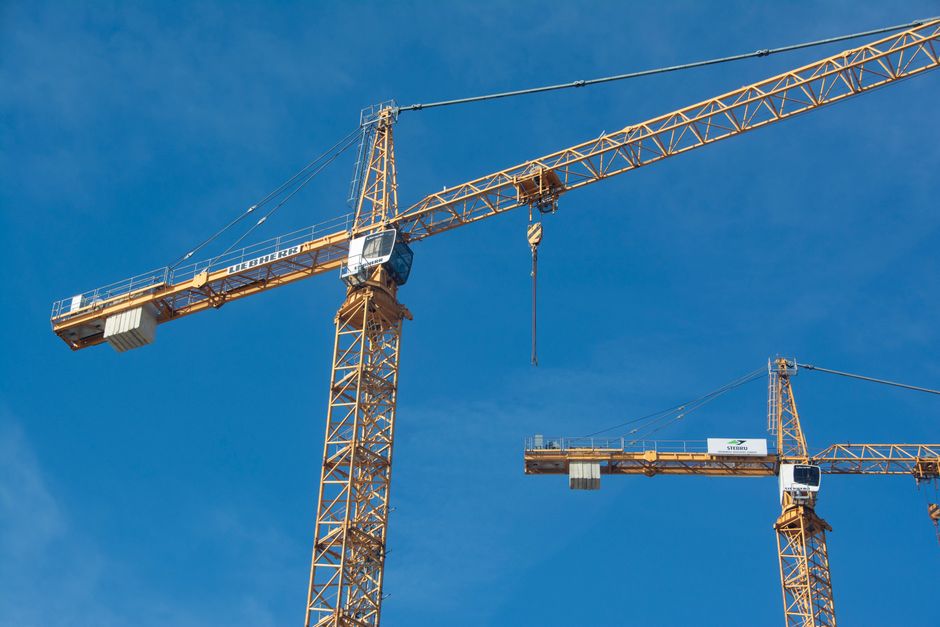 Imagem de duas torres de guindastes em um canteiro de obras sob céu claro, representando construção civil e infraestrutura.