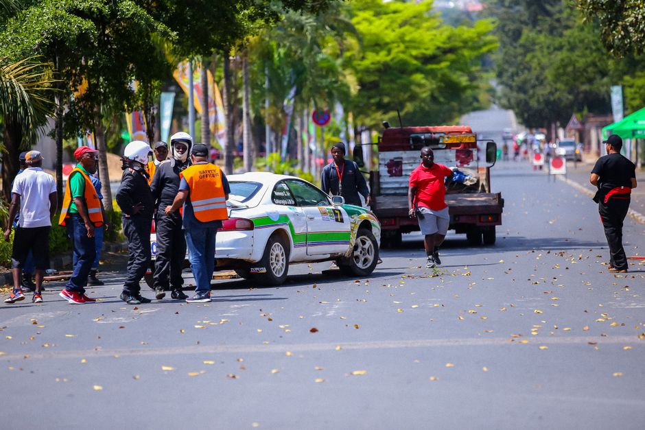 Imagem de uma cena de acidente de trânsito em uma rua movimentada, com várias pessoas, incluindo equipes de emergência e cidadãos, ao redor de um veículo policial e um caminhão de manutenção.