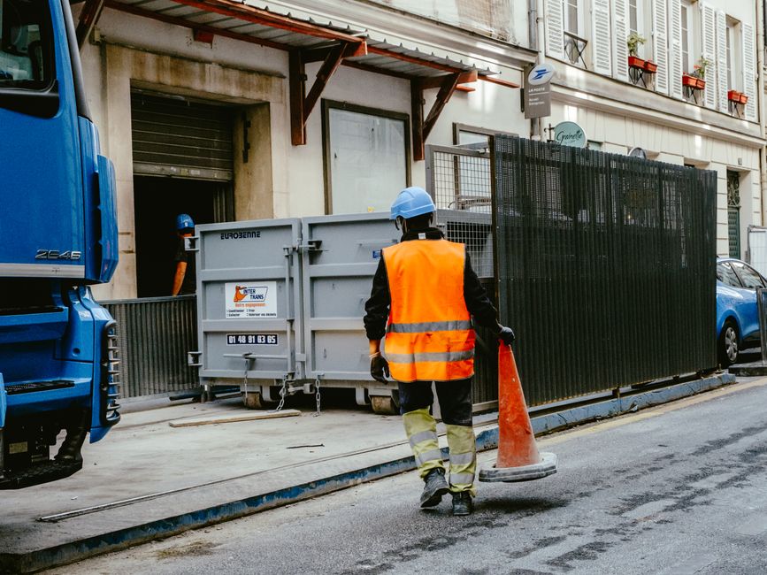 Funcionário de construção civil realizando sinalização de segurança na rua em frente a edifício residencial com escadaria e janelas com persianas.